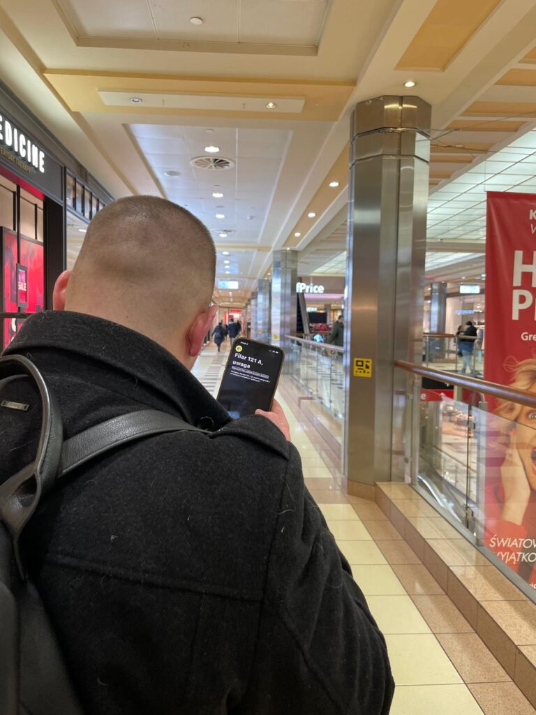 A person with a backpack uses the NaviWay app in the Sadyba Best Mall Passage. He holds the phone in front of him, and the screen shows navigation to the selected store.